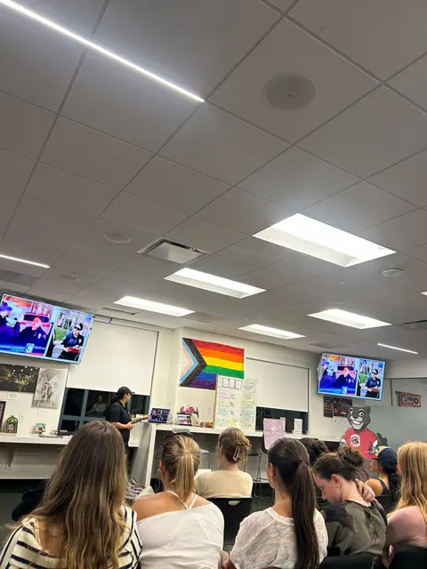 Students seated at a classroom meeting facing two presentation screens, a progress pride flag on the wall behind them.
