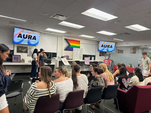 A full room of students at an AURA meeting, the club's name on screens at the front and a progress pride flag on the wall.