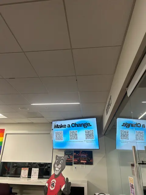 Classroom screens showing a slide reading "Make a Change" with a QR code beside student artwork on the wall.