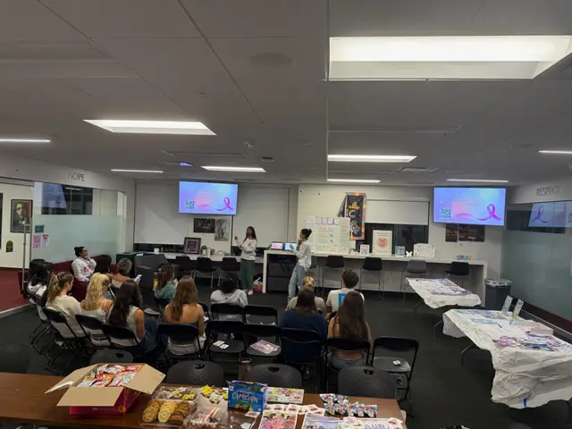 A meeting in progress with students seated in rows and a table of materials along the side of the room.