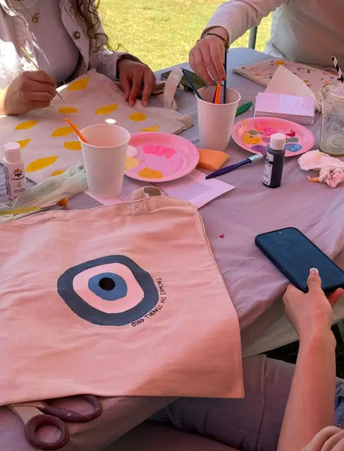 Hands painting an evil-eye design onto a canvas tote bag at a craft table.