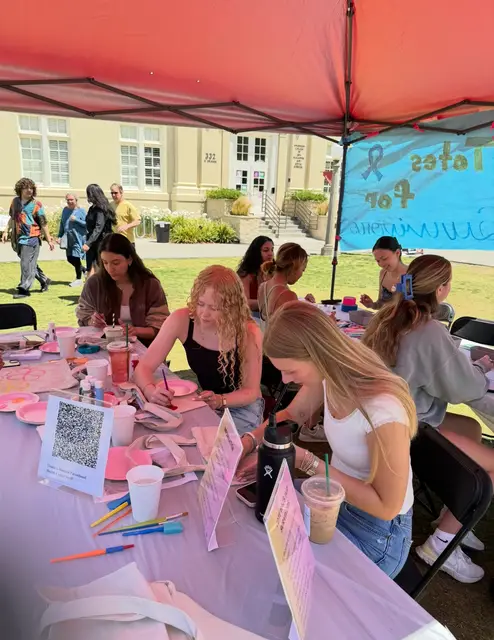 Students decorating tote bags together under a pink canopy at an outdoor event.