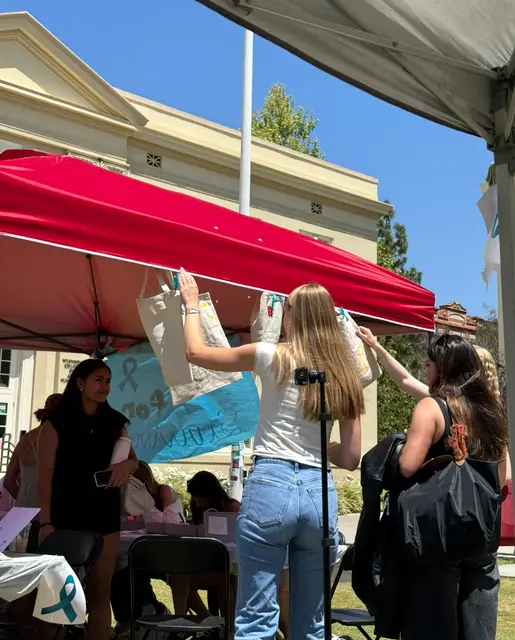 Students hanging a hand-painted banner at an outdoor tabling event under a red canopy.