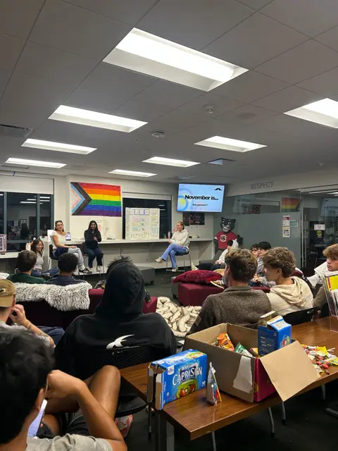 Students seated on lounge couches watching a presentation, a progress pride flag on the wall.