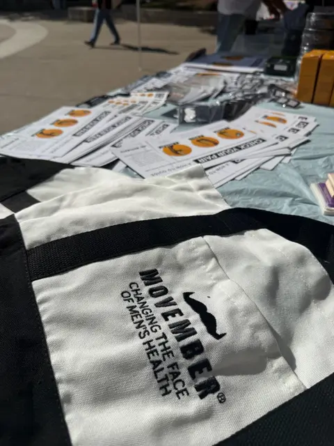 A Movember tote bag reading "Changing the face of men's health" resting on printed handouts.