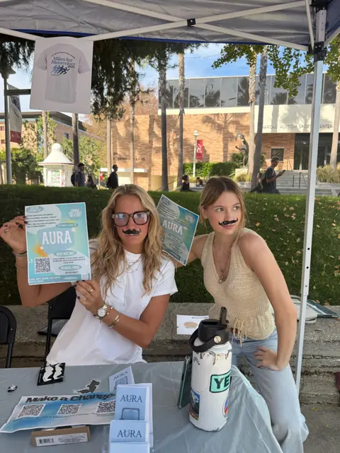 Two students with drawn-on moustaches holding AURA flyers at a Movember awareness table.