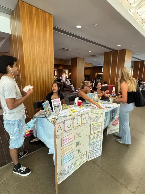 An indoor tabling setup with a hand-made AURA poster board and students gathered around it.
