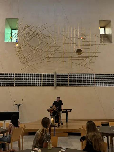 A performer on stage during an open-mic event, watched by a seated audience.