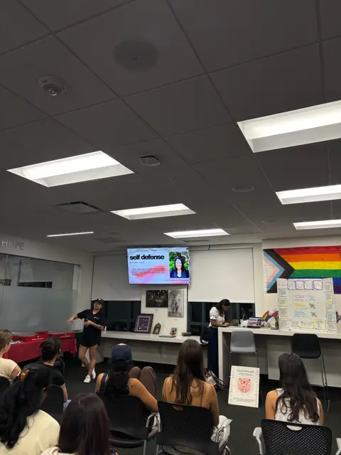 A presenter standing beside a screen reading "self defense" while students watch.