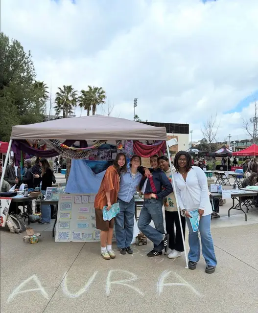 AURA members standing together at an outdoor campus fair behind their name chalked on the pavement.